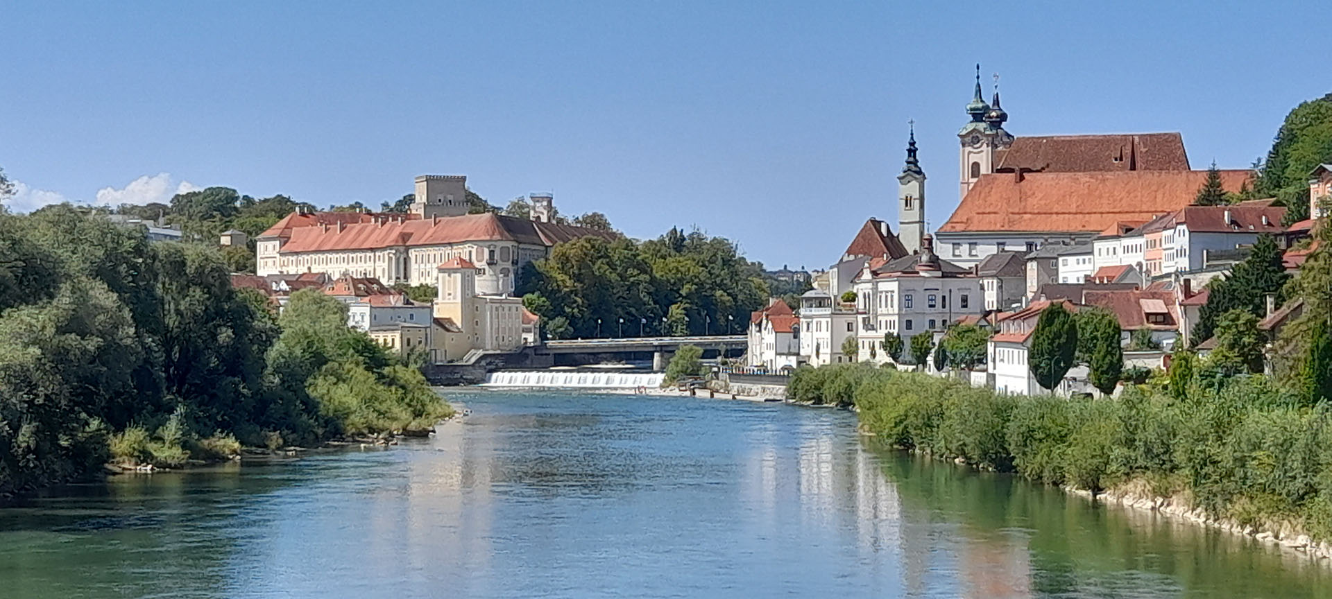 Blick auf die Stadt Steyr von einer Brücke mitten am Fluss, wir sehen Kirchtürme, Brücken, einen kleinen Wasserfall, ein Schloss mit Turm und Zinnen, links und rechts eingerahmt vom grünen Ufer